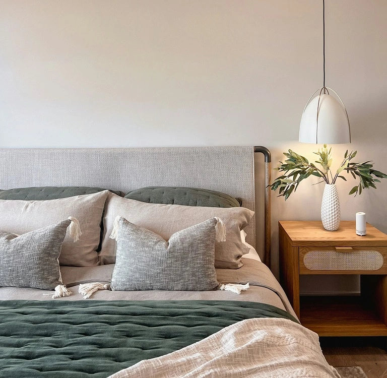 Modern bedroom with neatly arranged bedding in neutral tones, featuring layered pillows in different textures, a soft quilted blanket, and a wooden nightstand with a white vase and pendant lamp.