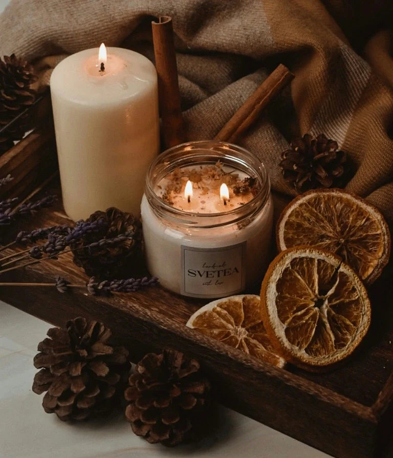 Cozy arrangement with a lit jar candle and pillar candle surrounded by pinecones, dried orange slices, cinnamon sticks, and lavender on a wooden tray.