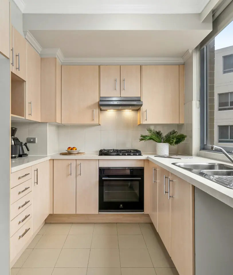 Modern kitchen with light wood cabinets, built-in oven, gas cooktop, white countertops, and a large window, decorated with a potted plant and fruit bowl.