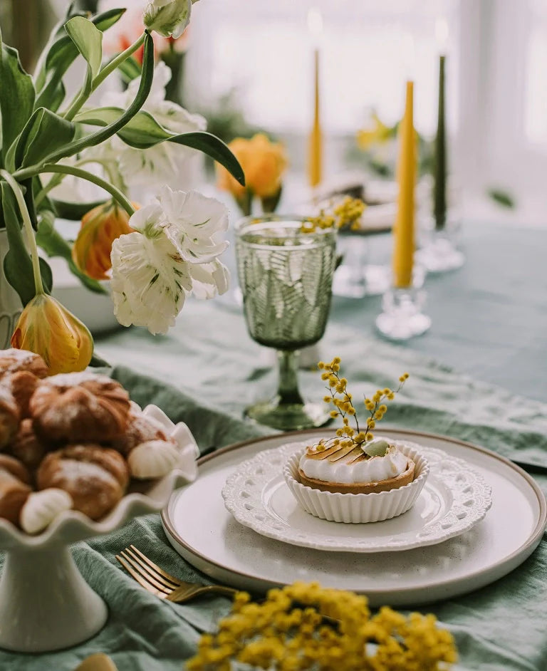 Elegant spring dining table setting with tulips, yellow candles, green glassware, pastries, and a decorated dessert on a white plate with lace-style detail.