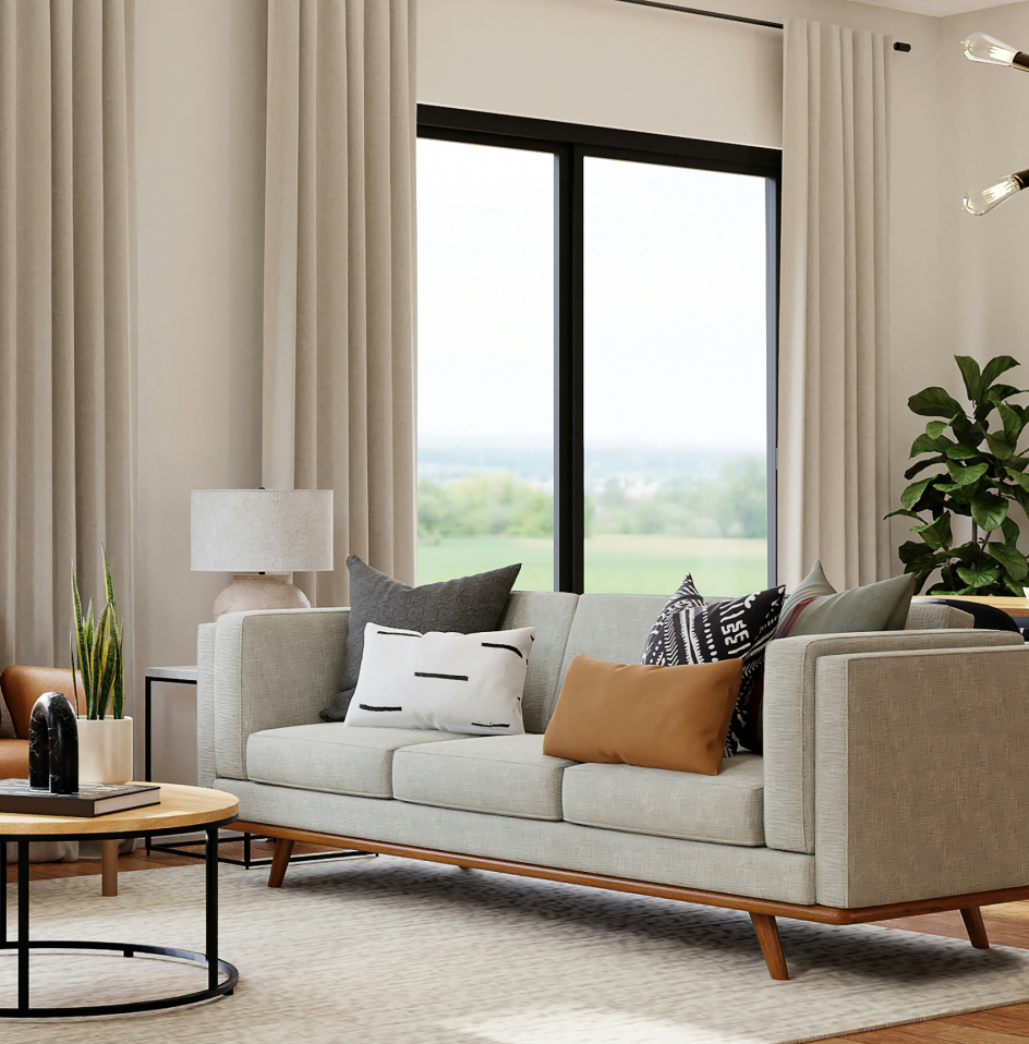 Modern living room with neutral décor, featuring a light gray sofa with assorted pillows, a round wooden coffee table, a table lamp, and a large window with beige curtains and a view of greenery outside.
