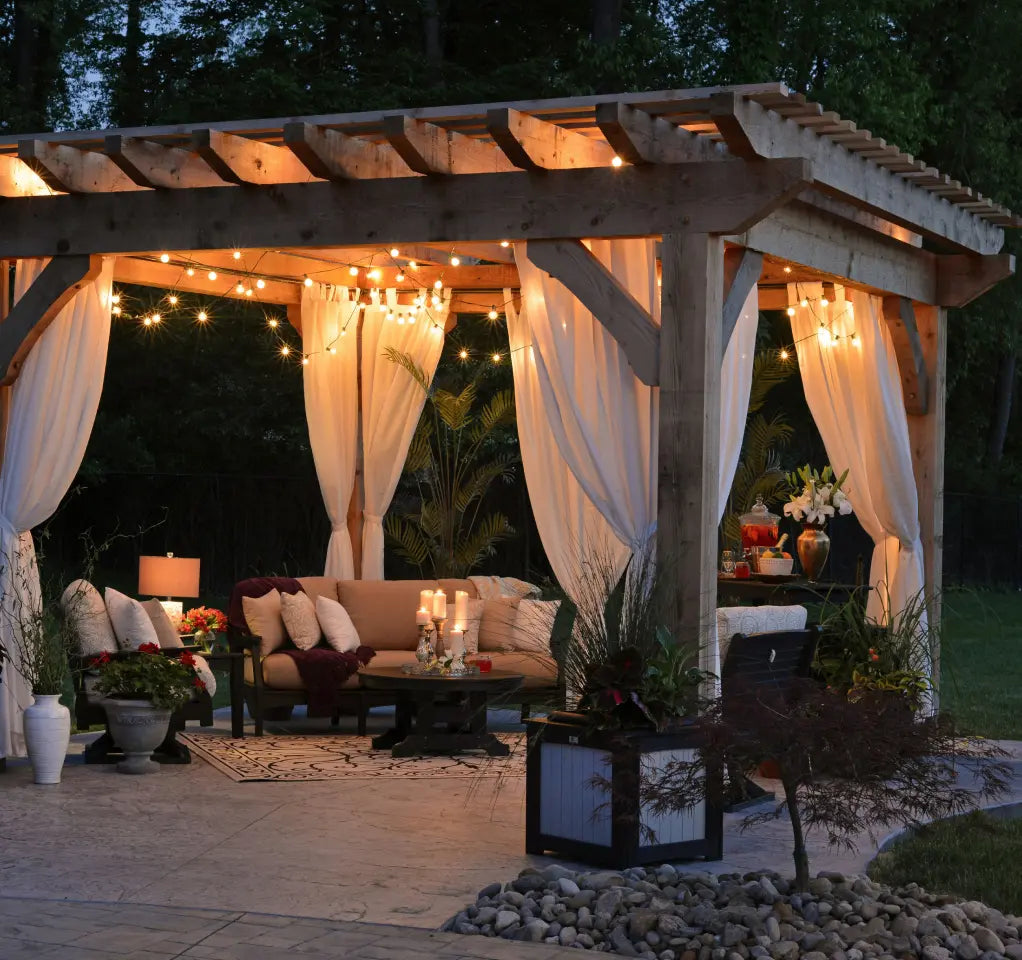 Outdoor patio with a wooden pergola draped in white curtains, string lights, and cozy seating with candles, plants, and decorative accents, creating a warm and inviting evening setting.