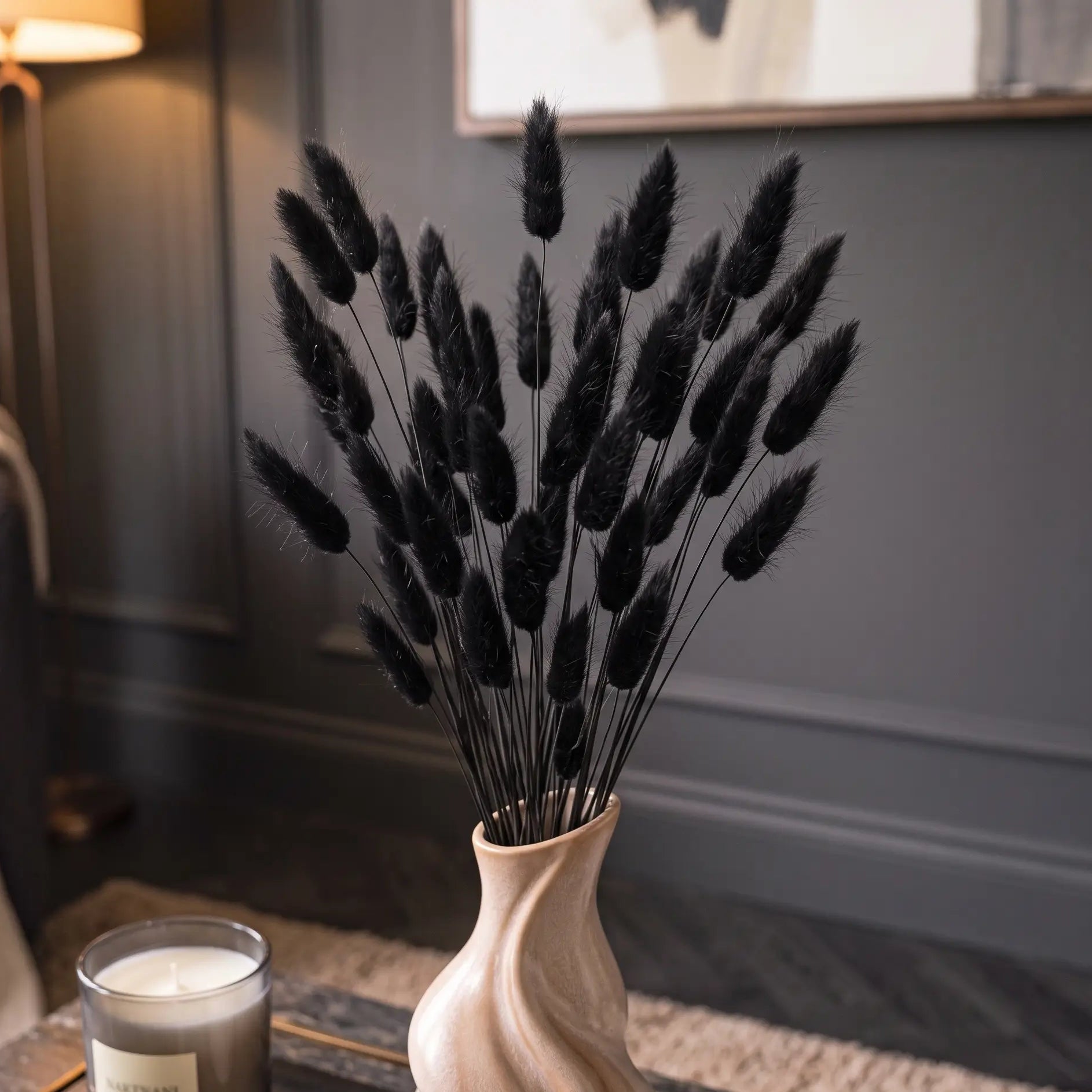 Black bunny tail grass dried flower stems arranged in a light beige ceramic swirl vase, set on a dark wooden table in a cozy room with a lit candle and a floor lamp in the background.