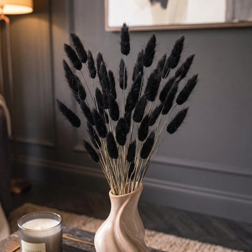 Black bunny tail grass dried flower stems arranged in a light beige ceramic swirl vase, set on a dark wooden table in a cozy room with a lit candle and a floor lamp in the background.