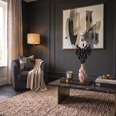 A styled living room corner with dark charcoal paneled walls, a large abstract painting, a sculptural vase filled with Black Dried Bunny Tails a gray armchair with a cream throw, a black coffee table, and a textured beige rug.