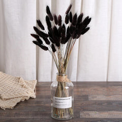 A clear glass bottle vase with a white label and twine neck wrap, filled with black dried bunny tail grass stems, set on a wooden tabletop against a white curtain backdrop.