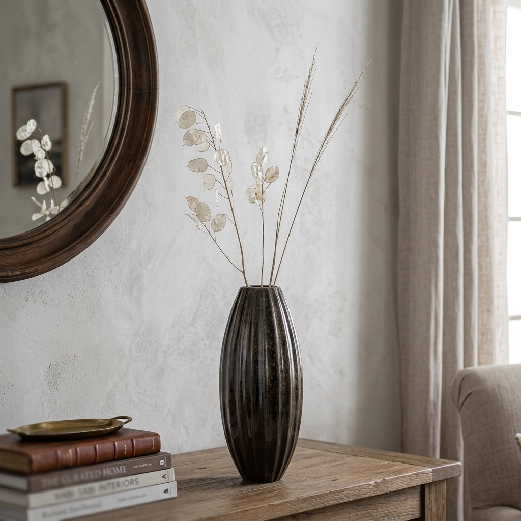 A dark brown ribbed ceramic vase filled with dried beige botanical stems, styled on a wooden console table in a living room setting next to a round mirror and curtains.
