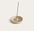 A round, shallow ceramic incense holder in a speckled beige stoneware finish, with a single incense stick standing vertically in the center, set against a solid off-white background.