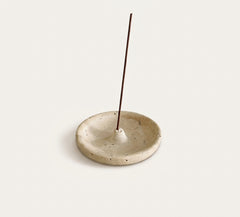 A round, shallow ceramic incense holder in a speckled beige stoneware finish, with a single incense stick standing vertically in the center, set against a solid off-white background.