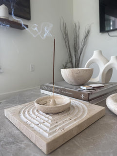 Incense burner with a single incense stick on a decorative stone tray, with books and a vase in the background.