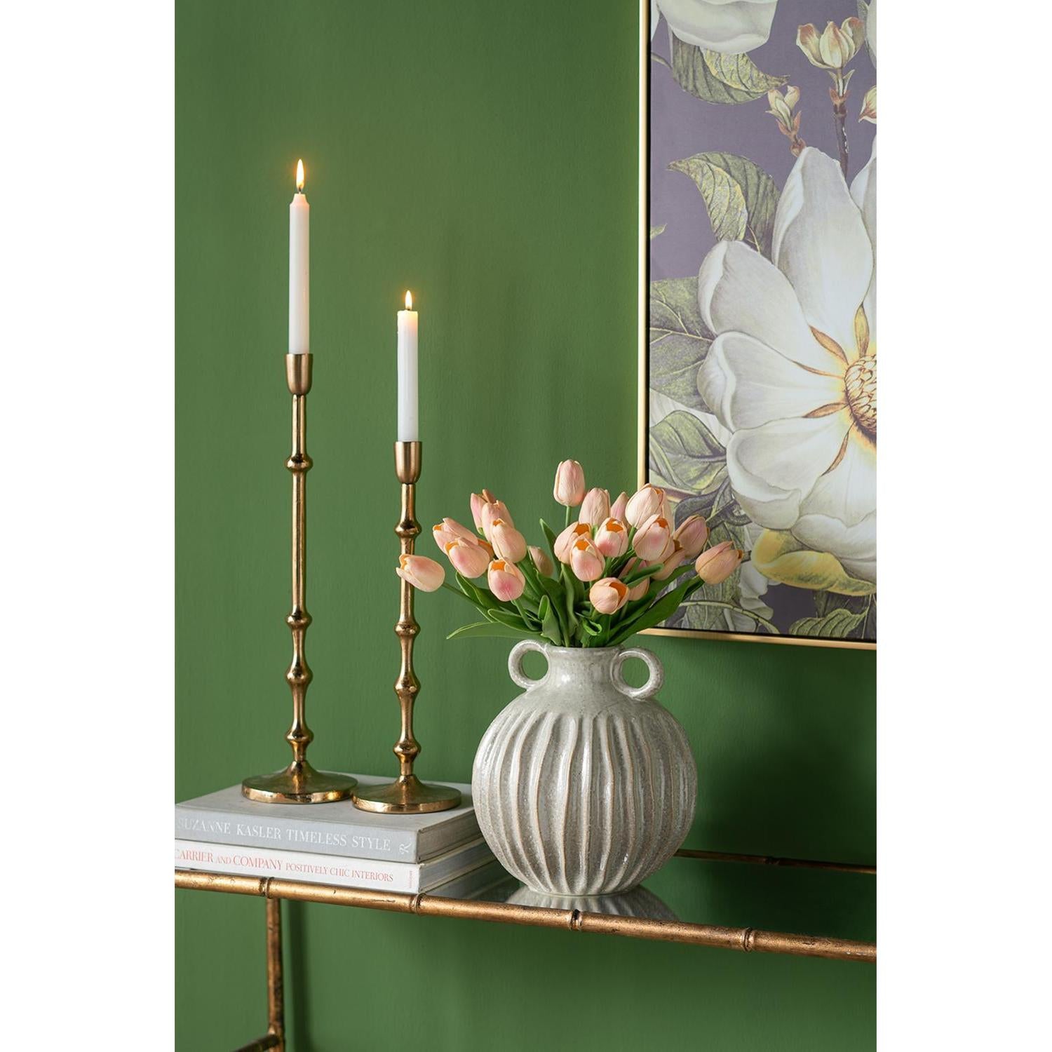 Decorative setup with candles in brass candlesticks and a vase of pink tulips against a green wall.