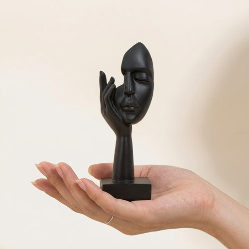 A matte black resin sculpture of a face resting on a hand, displayed on a person's open palm against a warm beige background.