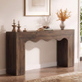 A brown wooden console table with a scalloped apron and blocky legs, styled with candles, books, and a vase in a bright living room.