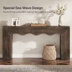 A rustic dark brown MDF console table with a scalloped apron detail, styled in a home entryway setting with decorative items, a vase of flowers, and a woven basket on a wood floor.