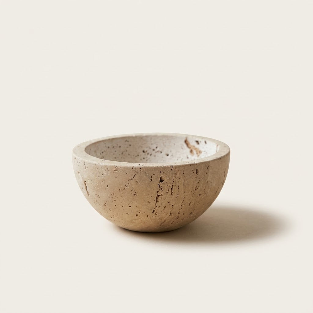 A small round travertine stone bowl with a textured, speckled beige surface, photographed against a solid off-white background.