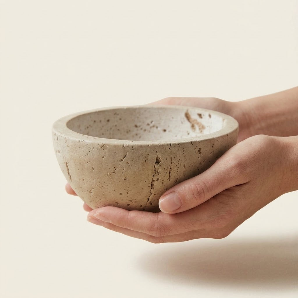 A person's hands holding a small, round concrete bowl with a speckled beige and gray texture against a solid cream background.