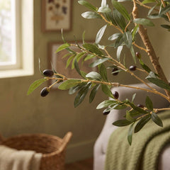 A detailed close-up of the artificial olive tree's branches, focusing on the slender, realistic green leaves and a sparse scattering of dark purple faux olives. The textured brown trunk and out-of-focus woven wall decor can be seen softly blurred in the background.
