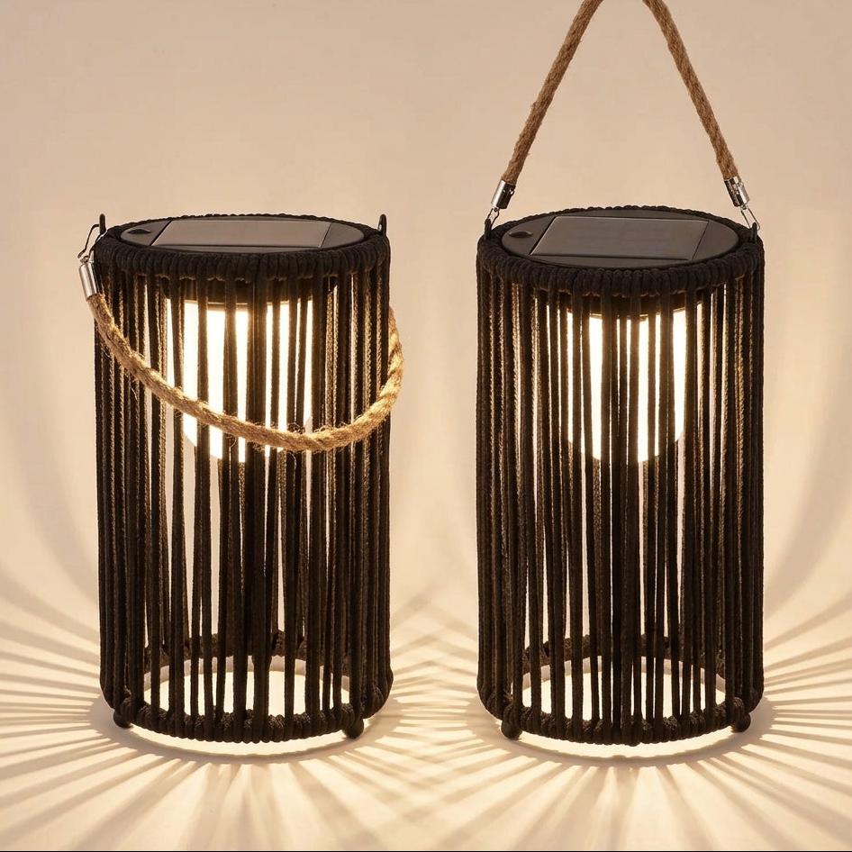 Two black rope lanterns with solar panel tops and rope handles, shown lit against a warm beige background with dramatic shadow rays.