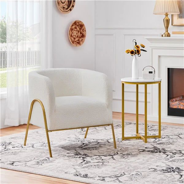 A modern ivory white upholstered accent chair with curved gold metal legs sits beside a small round gold side table in a bright living room setting.