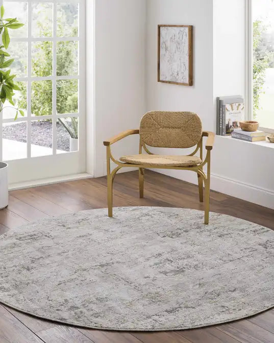 A round Meral Thick Luxe Boutique Area Rug with a distressed gray and white pattern, styled in a bright room with a wooden chair on top.