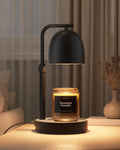 A modern black metal candle warmer table lamp with a dome-shaped shade hovers above a lit scented candle in a glass jar on a round base, placed on a tabletop in a softly lit room.