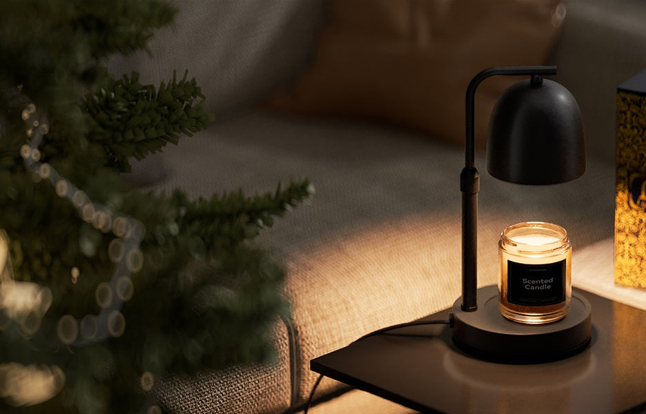 A scented candle in a glass jar sits on a Black Candle Warmer Lamp base next to a sofa in a cozy living room setting, with a blurred Christmas tree in the foreground.