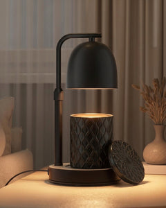 A modern black candle warmer lamp with a dome-shaped shade and the Forbidden black textured ceramic candle, shown lit on a tabletop in a softly styled interior setting.