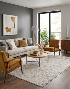 A styled living room featuring a light beige sofa, two mustard yellow armchairs, a round wooden coffee table, and a large distressed beige  Meral Thick Luxe Boutique Rug on dark wood flooring. The room has a gray accent wall with a framed abstract print, a floor lamp, and a large window looking out onto green plants.