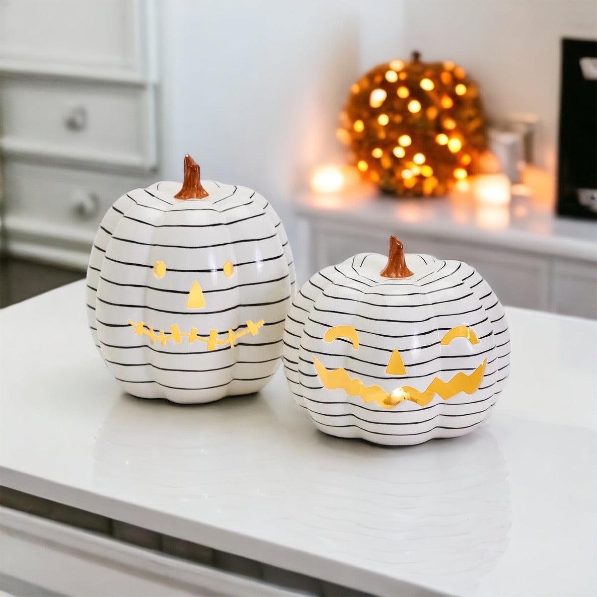 Two white pumpkins with black stripes and LED illuminated smiles on a white surface, with a blurred background of lights and furniture.