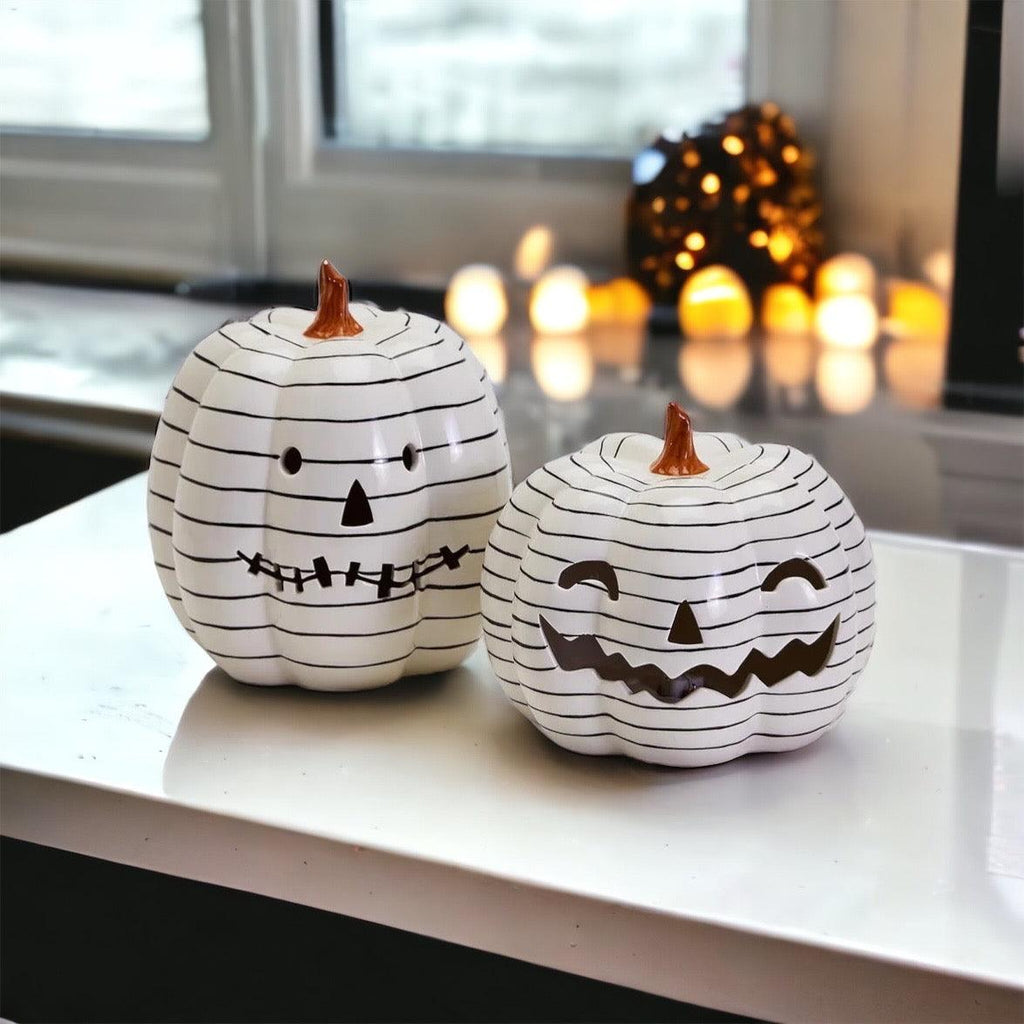 Two white pumpkins with black line art designs on a table with blurred lights in the background