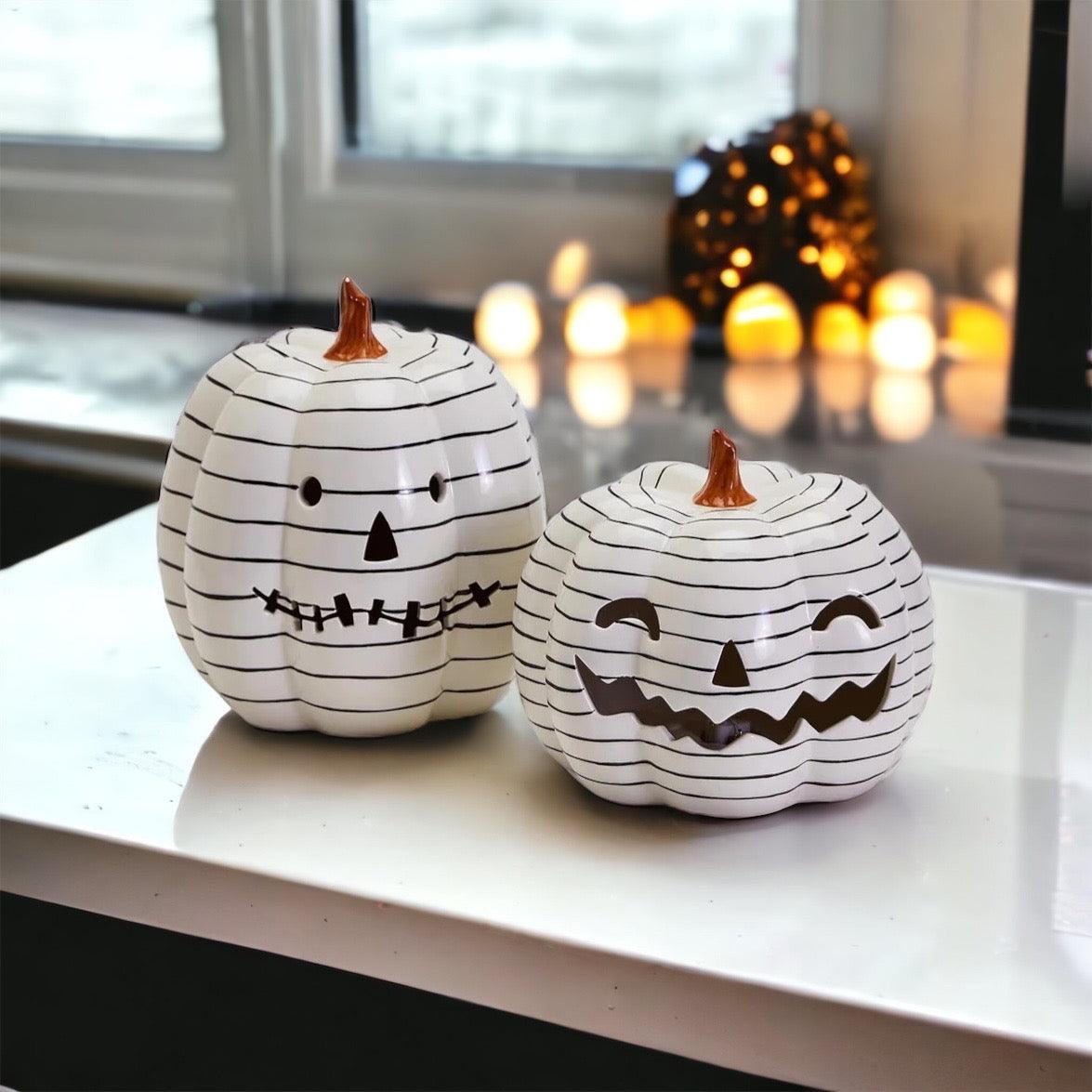 Two white pumpkins with black line art designs on a table with blurred lights in the background