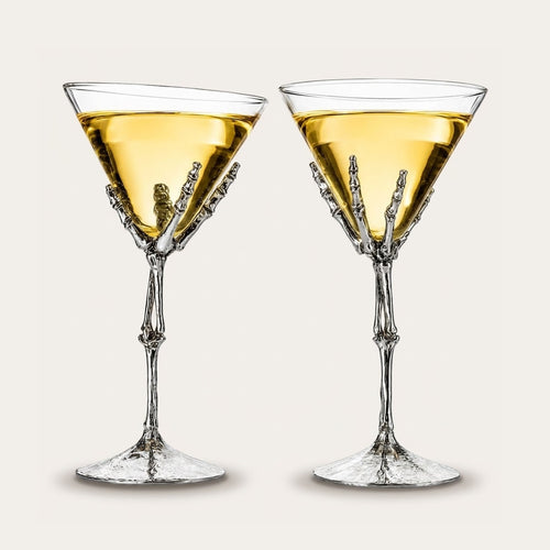 Two clear martini-style glasses with sculptural skeletal hand stems, each filled with a golden yellow drink, shown on a plain light beige background.