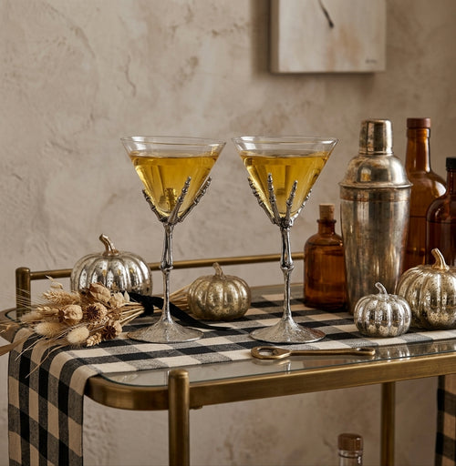 Two stemmed martini glasses filled with a yellow drink, styled on a bar cart with pumpkins, bottles, and a shaker against a textured beige wall.
