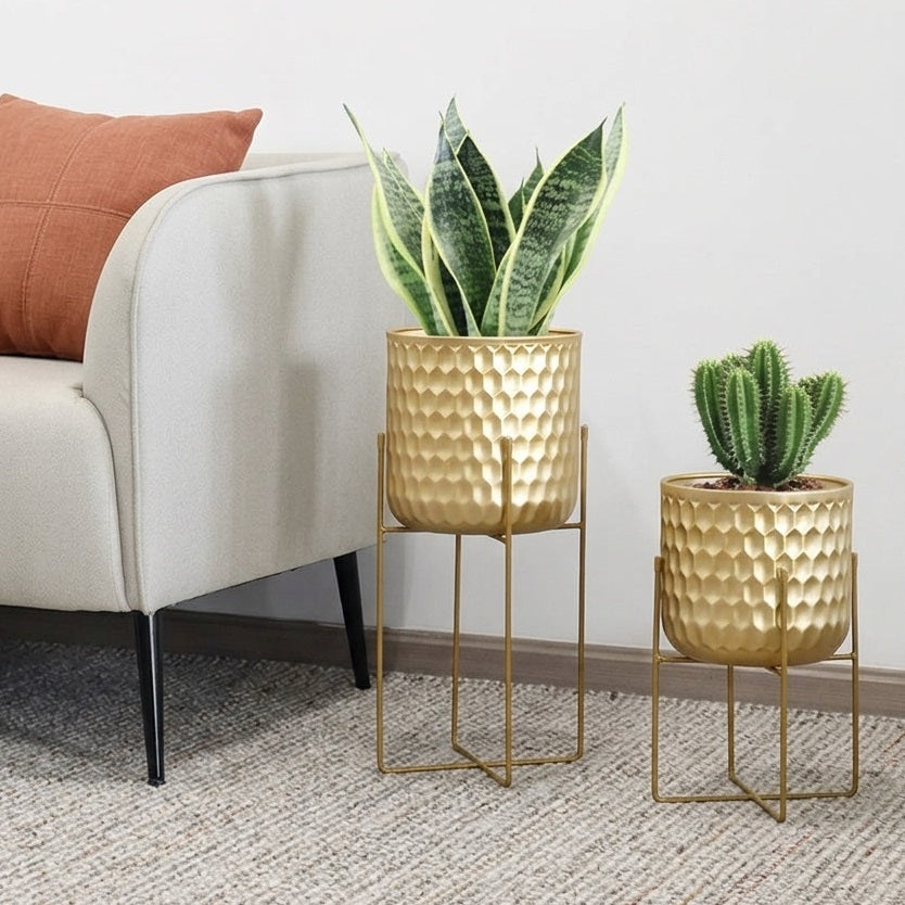 Two gold textured planter pots on slim gold metal stands, styled in a living room next to a light gray armchair with a rust-colored pillow. One pot contains a snake plant and the other a small cactus.