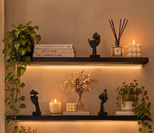 Two black floating shelves mounted on a beige wall, styled with books, black sculptural figurines, a reed diffuser, a lit candle, a bubble candle, and potted green plants. Warm LED under-shelf lighting illuminates the scene.